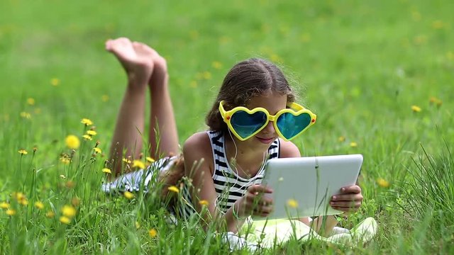 Beautiful girl in big sunglasses in shape of hearts with tablet computer lies on the grass. Girl playing the game on his tablet computer. Little girl in glasses lies on green grass and uses tablet pc