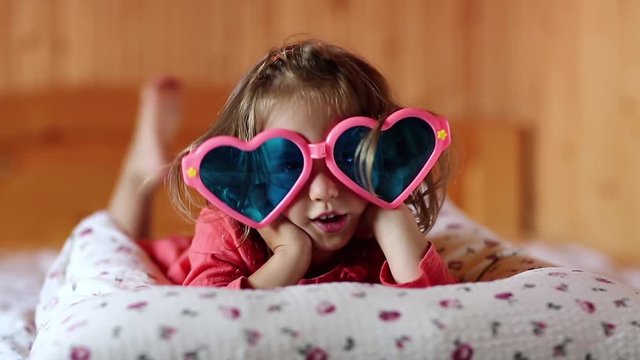 Little girl in big glasses in the shape of hearts lies on the bed. Funny little girl lies on the pillow and looks at the camera