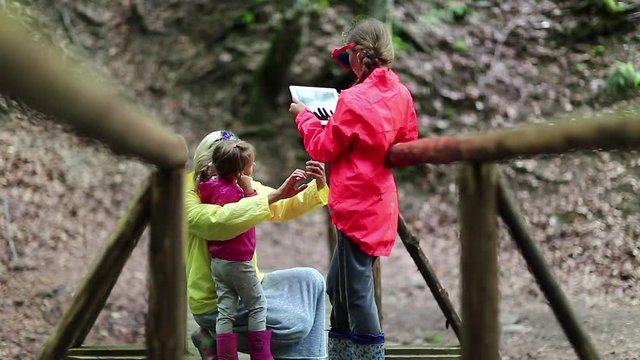 Family with electronic devices on the wooden bridge in the forest. Women in color jackets with tablet pc and smartphone. Mother and two daughters with smartphone and tablet computer on the nature