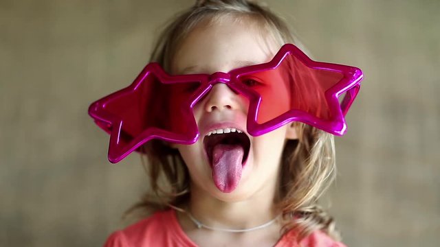 Funny little girl in pink glasses looks at the camera. Pretty little girl in big glasses in the shape of stars looks at the camera