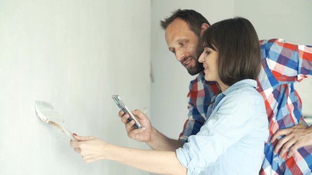 Young couple with smartphone talking about project while painting wall at their new home 
