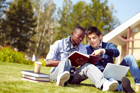 Two young students sitting on grass in campus and looking through a textbook