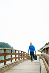 Strong sportsman arriving at his outdoor training spot on a cloudy autumn day with a sport bag on a wooden walkway. Fitness sporty man in Rodiles, Asturias.