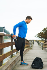 Fit sports man doing stretches before an outdoor running training on a cloudy autumn day with a sport bag on a wooden walkway. Fitness sporty man in Rodiles, Asturias.