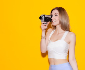 Beautiful fashionable girl posing and holding a vintage movie camera in a top and a skirt on a yellow background in the studio. Gorgeous Woman Portrait. Sexy Glamour Girl on Yellow background
