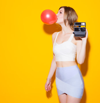 Fashionable modern girl posing in colorful top and skirt inflates the red bubble from chewing gum and with a vintage camera in her hand on yellow background in the studio. Fashion Beauty Girl. 