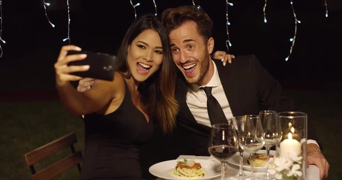 Beautiful romantic couple enjoying an elegant dinner in a restaurant posing for a selfie smiling into the camera on their mobile phone