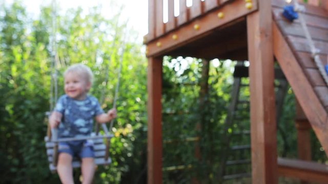 One years-old baby boy with blue eyes play on the swing outdoor on a sunny day. Mum helps the little son swinging on the swing. slowmotion