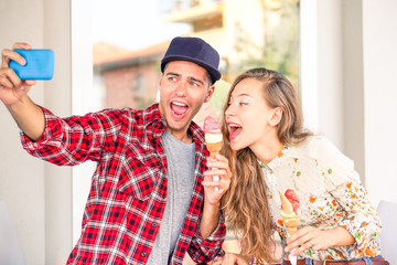 Playful couple eating ice cream taking funny selfie outside shop - Girlfriend teasing boyfriend with funny face in self photo on joyful holiday moment - Concept of friends having fun using technology