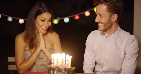 Pretty young woman celebrating her birthday blowing out the candles on the cake at a party watched by her loving boyfriend of husband