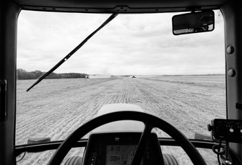 A tractor window framing a field of wheat being harvested by two combines under cloudy rural black and white landscape