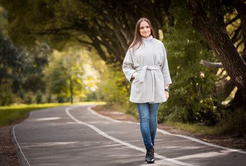 Attractive confident young woman enjoying her time outdoors in park. Wearing coat, Walking with happy smile. Fall landscape. Yellow leaves on trees. Healthy lifestyle
