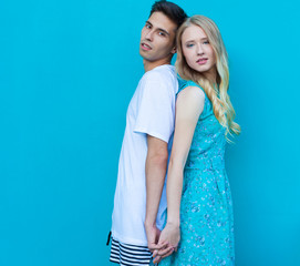 Stunning sensual outdoor portrait of young stylish fashion couple posing in summer. Guy hugging girl from behind, stand back to back. Hispanic man, Caucasian girl. Bright blue green background