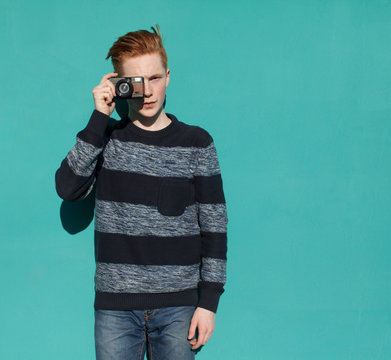 Young redhead man in a sweater and jeans standing next to turquoise wall and taking photos vintage camera warm summer sunny day