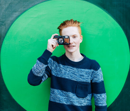 Young redhead man in a sweater and jeans standing next to green wall and taking photos vintage camera