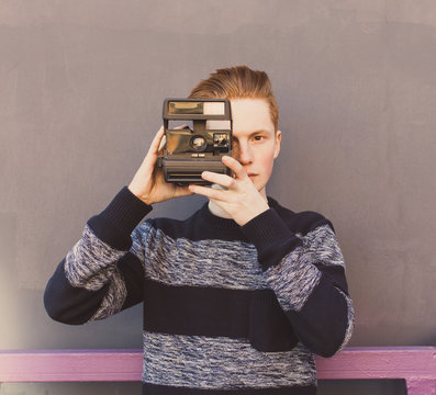 Young redhead man in a sweater and jeans standing next to gray wall and taking photos vintage camera warm summer sunny day