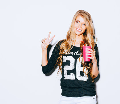 Happy Young Woman Showing Victory Sign and holding a pink glass with straw in hand On White Background. Indoor. Warm color.