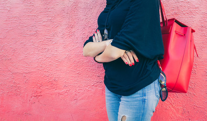 Fashionable beautiful big red backpack on the arm of the girl in a fashionable jeans, posing near the pink wall on a warm summer night. Part of body. Warm color