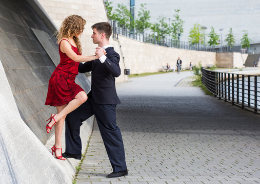 two young people - a man in black suit and a woman wearing red dress - dancing tango outside under the bridge