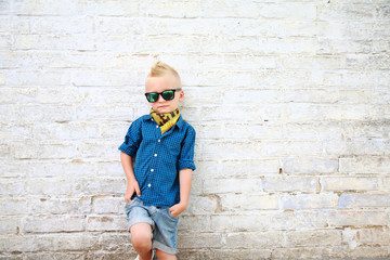 стильный мальчик в солнцезащитных очках возле кирпичной белой стены - obrazy, fototapety, plakaty