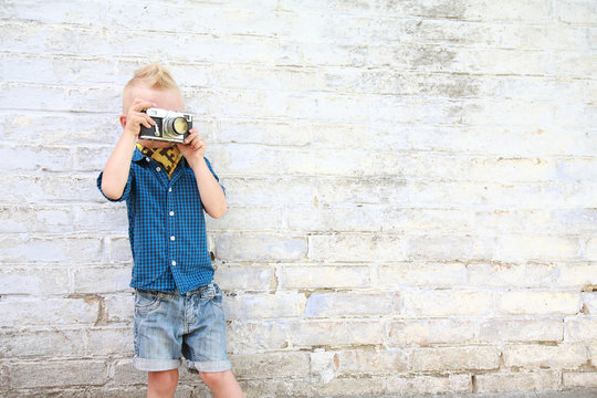 мальчик блондин с фотоаппаратом возле белой кирпичной стены 