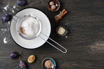 Plum pie cooking process. Powdered sugar, plum fruits and spices on dark wooden background.