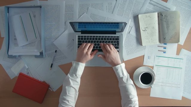 Top view businessman working at office desk with his laptop drinks coffee and spills it on a piece of paper rumples it and throws away surrounded by a lot of paperwork and financial reports, notebooks