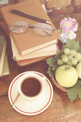 Cup of fresh tasty coffee on a wooden table, vintage books, fresh fruits and vase with pink flower. Romantic, retro decor elements. Old brick wall background