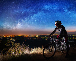 Young cyclist with mountain bike on top of the hill observing the night city view