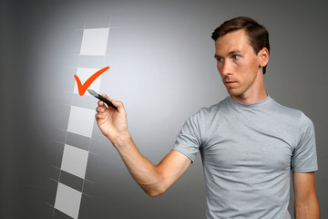Young man in t-shirt checking on checklist box. Gray background.