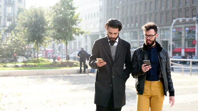 Half length of two young handsome caucasian bearded brown hair businessmen walking in the city, talking to each other an discussing using smart phone- business, technology, work concept