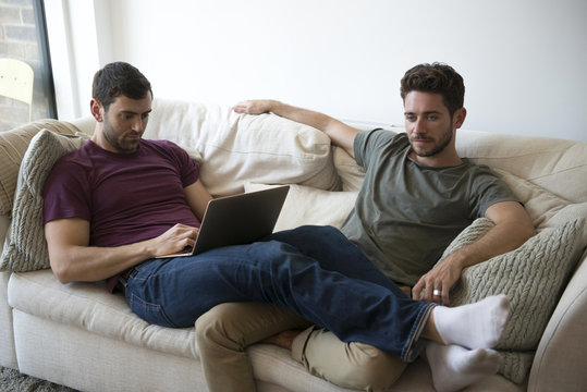 Gay Male Couple Relax On Sofa Watching TV And Using Laptop