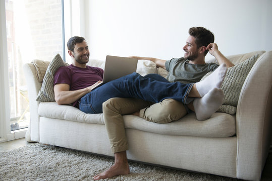 Gay Male Couple Relax On Sofa Watching TV And Using Laptop