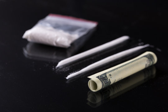 Rolled hundred dollars banknote, two lines and plastic packet of cocaine on black background, closeup 