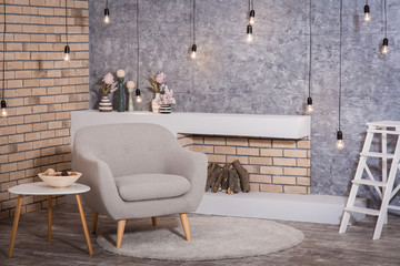Loft style interior: modern fireplace with firewood, cozy armchair and coffee table. Garland of bulbs hanging from the ceiling. 