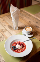 Serving table with white milky creamy pudding with jam and berries blueberries, strawberry, raspberry on a white plate and cup of coffee in cafe or restaurant