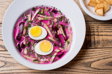 Close-up of cold beetroot soup with cabbage and egg served with fried potatoes on a white plates at the rustic wooden background