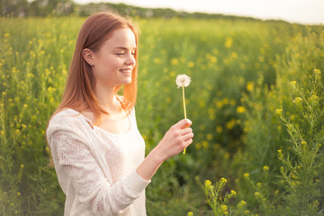 Young spring fashion woman blowing dandelion in spring garden. Springtime. Trendy girl at sunset in spring landscape background