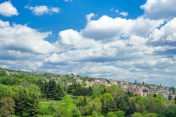 Fototapeta premium City landscape. The town, surrounded by greenery on the background of large volumetric clouds. Cottage village, located on a hillside. Trees in bright spring foliage