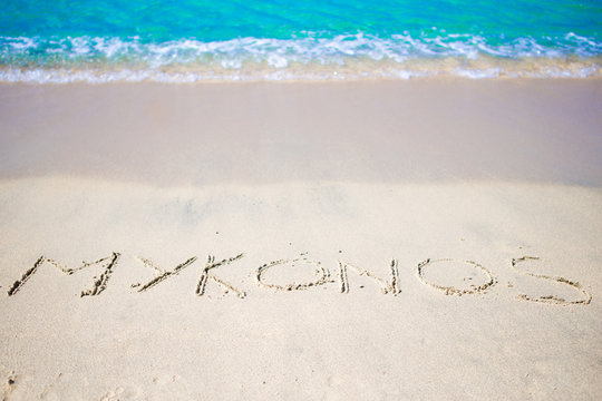 Word Mykonos handwritten on sandy beach with soft ocean wave on background