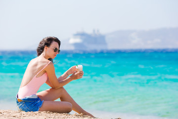 Young beautiful woman with coffee on the beach during tropical vacation. Girl enjoy her wekeend on one of the beautiful beaches in Mykonos, Greece, Europe.