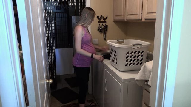 A young expecting woman places dirty clothes and towels into the washer.  	