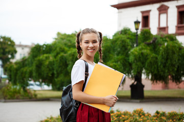 Young beautiful cheerful female student smiling, holding folders outdoors, park background.