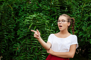 Surprised young beautiful female student in glasses pointing finger away, leaves background.