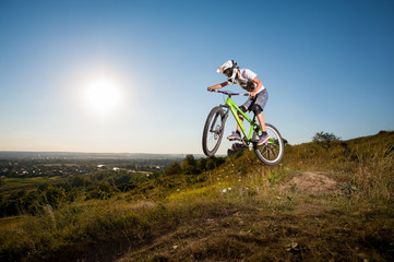 Obraz premium Man making extreme jump on a mountain bike on the hill against blue sky, sun and greenery into the distance. Cyclist is wearing white sportswear helmet and glasses.