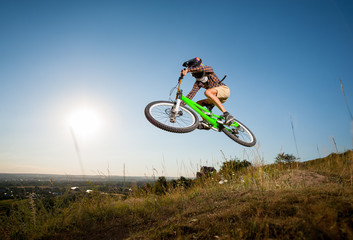 Guy high jump on a mountain bike on the hill against blue sky, bright sun, greenery and small town...
