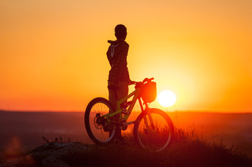 Fototapeta premium Silhouette of the guy with the bike at the top of mountain on the sunset background. Cyclist looking to the evening bright sun
