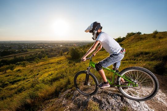 Cyclist in helmet and glasses on mountain bicycle stands on the precipice of slope and looking down under blue sky