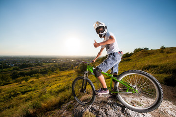 Fototapeta na wymiar Bicyclist in helmet and glasses on mountain bike stands on the precipice of hill and showing thumb up gesture of good class under blue sky and sun. Wide angle view