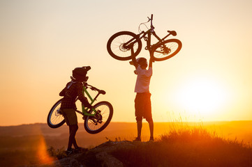 Fototapeta na wymiar Two guys in helmets holding a mountain bikes in hands on the top of the mountain against evening sky with bright sun at the sunset